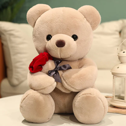 Beige teddy bear with a red rose and purple bow on a soft surface.